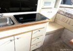 Compact camper kitchenette with a stainless sink, black glass stovetop, and light wood trim; cream cabinets and drawers beneath, and a bed area visible to the right.