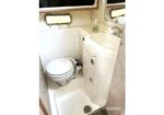 Compact boat bathroom with a toilet beside a small sink and soap dispenser; windowed walls and a drainage floor visible.