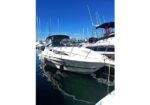 White motorboat docked at a marina, with several sailboats and tall masts in the background and calm water reflecting the hulls.