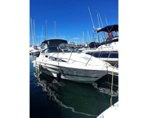 White motorboat docked at a marina, with several sailboats and tall masts in the background and calm water reflecting the hulls.