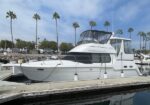 White motor yacht docked at a marina with palm trees and blue sky in the background; boat tied to a wooden pier.
