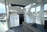 A white yacht cockpit area with seating, stairs, and a cabinet on a carpeted deck, with water and marina visible outside.
