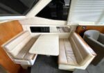 U-shaped beige leather seating surrounding a speckled rectangular table in a yacht cabin, with wood paneling and windows nearby.