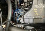 Close-up of an engine compartment showing blue ignition wires, black hoses, and a metal engine cover with a yellow emblem.