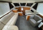 Boat salon interior with cream leather sofa, gray armchair, a table, wooden cabinets, and large windows with blinds.