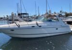White motor yacht docked at a marina with sailboats and masts in the background on a sunny day.