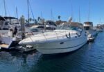 White motor yacht docked at a marina with other boats and palm trees in the background.