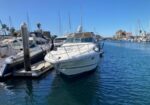 White motor yacht docked at a sunny marina with calm blue water and other boats in the background.