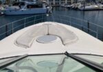 Bow of a white yacht approaching a marina with moored boats and calm water in the background, railing on sides visible.