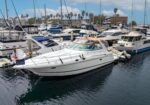 White motor yacht docked at a marina with other boats and palm trees in the background under a blue sky.