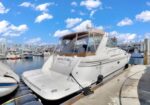 White motor yacht tied to a concrete dock at a marina, with calm water, other boats moored, and a bright blue sky with fluffy clouds.