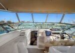 Luxurious white yacht cockpit with L-shaped seating, helm, navigation screens, and large windows overlooking a marina.