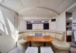 Cream curved boat-cabin seating around a wooden table, with overhead cabinets and two small porthole windows.