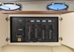 Boat electrical control panel with numerous switches and meters inside a cabinet, two round portholes above.