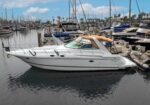 White motor yacht moored in a marina, with sailboats and palm trees in the background under a blue sky.
