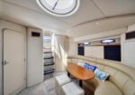 Cream yacht cabin with an oval wooden table, curved sofa seating, blue cushions, porthole windows, and stairs to the deck.