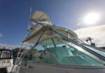 White motorboat with a raised cockpit and curved glass windshield docked at a sunny marina, blue sky overhead and sun glare visible on the glass.