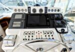 Helm of a boat with a steering wheel, multiple gauges, screens, and control panels arranged in a central console under large windows.