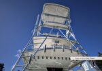 Tall tuna tower on a fishing boat with a metal ladder and seating, against a blue sky.