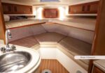 Interior of a boat cabin: tan U-shaped seating around a wooden floor, sink at left, overhead cabinets; forward bunks indicate it sleeps 4.