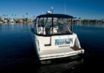 Rear view of a white motorboat on calm water, with a blue sign reading 'BEACH' and 'Long Beach, CA' on the stern; shoreline homes and palm trees in the background.
