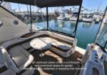 Luxurious yacht aft deck seating with curved white cushions around a round table, open cockpit overlooking a marina with boats and palm trees in the distance. Best spot for guests to relax underway or at anchor.