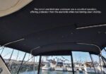 Boat cockpit with a dark bimini; a white label on the canopy reads: 'The bimini and Makrolon enclosure are in excellent condition, offering protection from the elements while maintaining clear visibility.' A marina with moored boats is visible beyond.