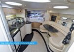 Interior of a yacht: compact galley on the left, curved beige seating with a small table on the right, bed area visible in back.