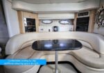 Yacht cabin lounge with cream curved seating around a black speckled table; built-in cabinets and porthole windows behind, showing comfortable day seating that can convert to sleeping space.