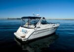 White motorboat with a dark canopy on calm blue water, distant shoreline visible under a clear sky.