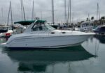 White motor yacht docked in a marina with sailboats in the background and a green canvas canopy overhead.
