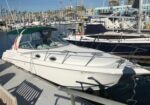 White motor yacht docked at a sunny marina with other boats and masts in the background