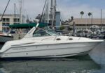 White motor yacht moored in a marina with a green canvas canopy; masts of other sailboats and buildings in the background.