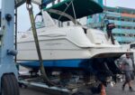 White motorboat lifted on a cradle in a marina boatyard, with a blue hull visible underneath and a man walking nearby.