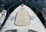 Top-down view of a white motorboat bow with a beige sunpad, stainless rails, and deck hatches, docked at a marina with calm water nearby.