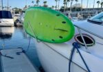 Bright green boogie board resting on the side of a white boat at a marina. Palm trees are in the background.