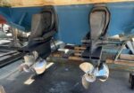 Rear view of a blue boat on a trailer, showing two stainless steel propellers and stern drive units in a boatyard.
