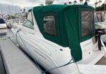 White motorboat with a green canvas canopy, docked at a marina; stern view with mooring lines and outboard motor visible under cloudy skies.