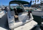 Rear view of a white motorboat at a marina, with a green canvas bimini, swim platform, and an outboard motor mounted at the stern among sailboats.
