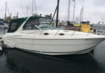 White motorboat docked at a marina with a green canvas cover and neighboring boats in the background
