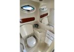 Compact boat bathroom (head) with white fixtures: toilet, small sink, oval porthole window, and wood trim; towels hang on the wall rack.
