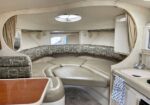 Curved beige leather seating arranged in a boat cabin with a central sunken floor, porthole window overhead, and a compact kitchenette to the right. Curtains line the walls behind the seating.