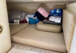 Under-seat storage area with beige cushions, folded blankets, a pink towel, and a small boxed item inside a vehicle.