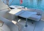 White boat deck with a U-shaped cushioned seating area and a foldable table on two pedestals; blue water in the background and a small American flag.