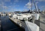 White motorboat docked at a marina with sailboats in the background and a walkway on the right.