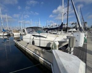 White motorboat docked at a marina with sailboats in the background and a walkway on the right.