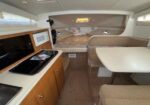 Interior of a small boat cabin: kitchenette with microwave and stove on left, sink, wooden cabinets, and a dining area with a table and cushioned seating on the right; bed at the bow with pillows visible.