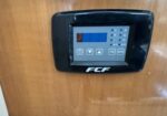 Digital thermostat mounted on a wooden wall, with a blue-backlit display and multiple control buttons.