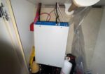 White electrical control box with a blue top mounted inside a cabinet, connected to red and black wires with plumbing items nearby.