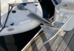 Close-up of a boat's anchor roller and mounted anchor on a dock, with water nearby and deck hardware visible behind.